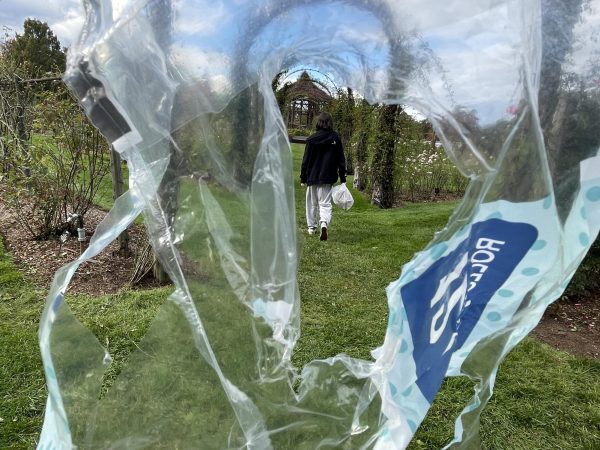 With the lens partially obscured by a plastic wrapping, Hall Records editor, Raina, walks through Elizabeth Park with a plastic bag of trash in hand as she approaches a disposal bin.