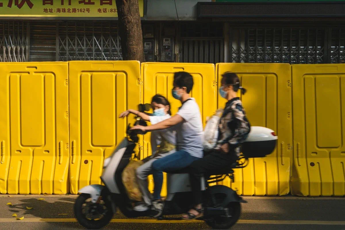 Taken in 2022 during the COVID lockdown in Guangzhou, a family of three wearing masks rides past on an electric scooter, as nearby shops are blocked off by yellow water-filled barricades.